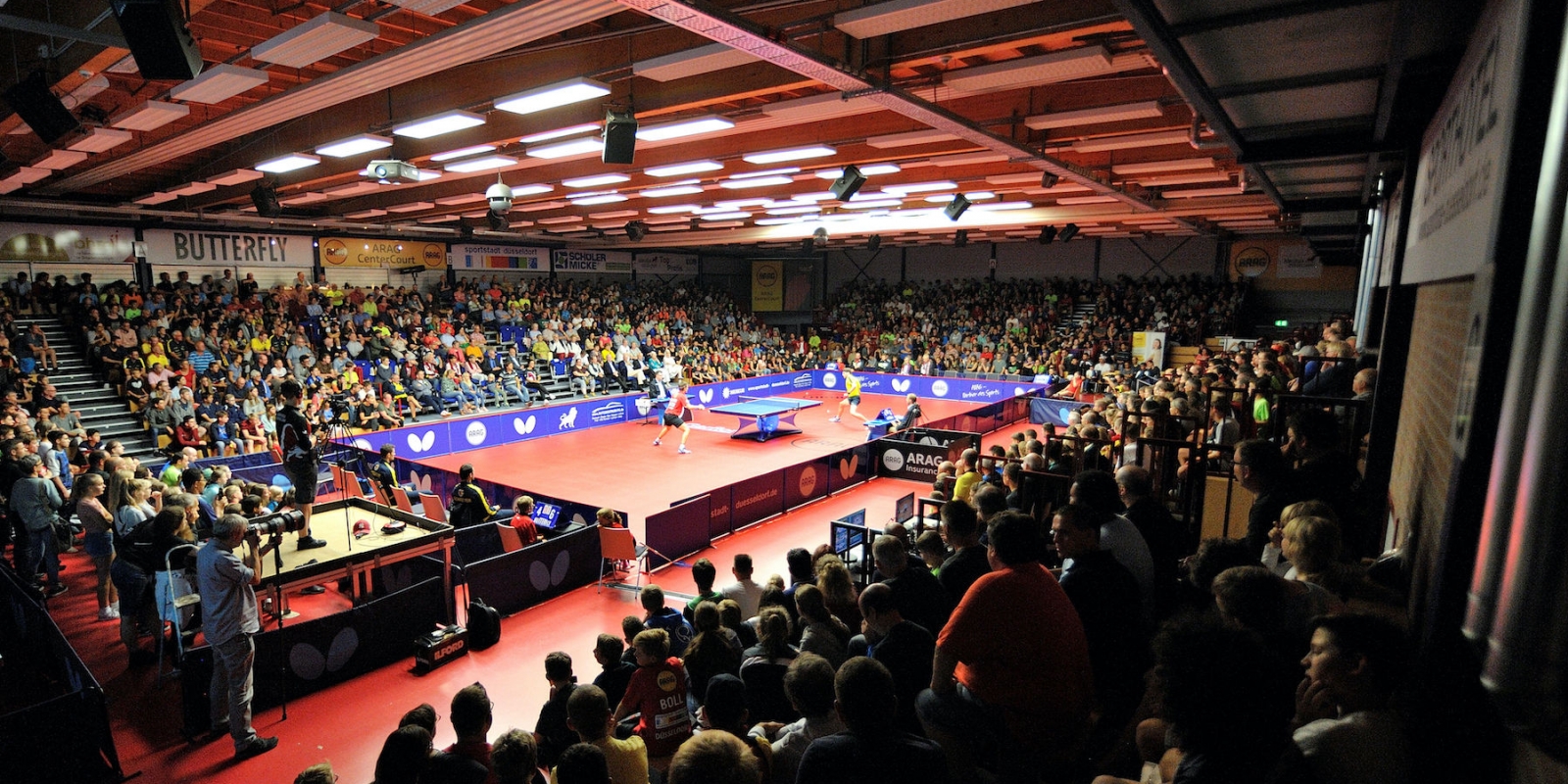 Tischtennis Halle mit vollen Zuschauerrängen. In der Mitte des Feldes spielen zwei Tischtennisspieler an einer Tischtennisplatte.